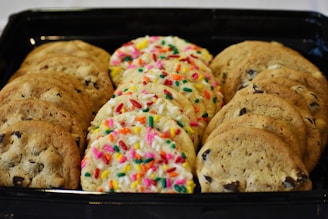 An assortment of cookies neatly arranged in a black tray, featuring chocolate chip cookies on both sides and a row of sugar cookies with colorful sprinkles in the center.