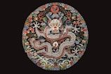 A detailed and vibrant embroidery showcases a mythical dragon surrounded by intricate floral and cloud motifs. The dragon is centrally featured with a fierce expression, and its scaled body winds through the circular design. The elaborate use of colors and textures lends an ornate and traditional feel.