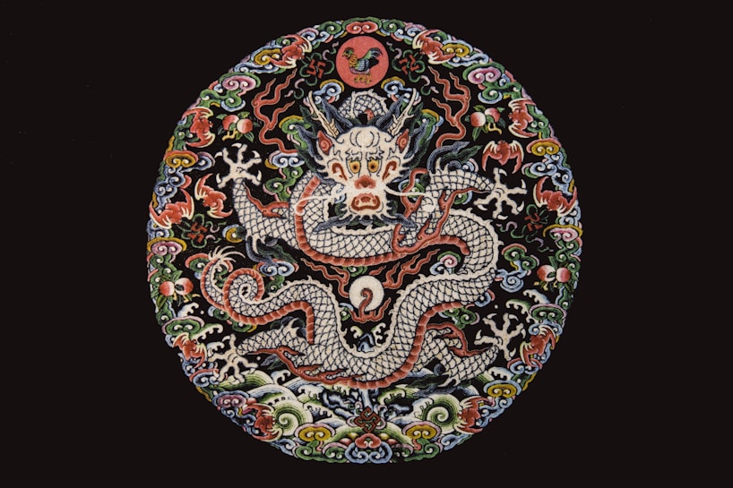 A detailed and vibrant embroidery showcases a mythical dragon surrounded by intricate floral and cloud motifs. The dragon is centrally featured with a fierce expression, and its scaled body winds through the circular design. The elaborate use of colors and textures lends an ornate and traditional feel.