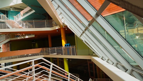 An interior view of a modern building featuring multiple levels with intersecting staircases and walkways. The architecture includes large glass panels allowing natural light to flood the space, creating an interplay of light and shadows. The color palette is predominantly warm, with orange, green, and brown elements.