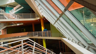 An interior view of a modern building featuring multiple levels with intersecting staircases and walkways. The architecture includes large glass panels allowing natural light to flood the space, creating an interplay of light and shadows. The color palette is predominantly warm, with orange, green, and brown elements.