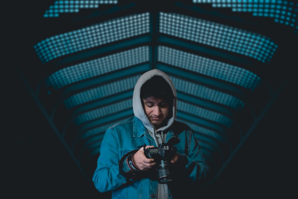 man with blue hoodie holding black Canon DSLR in dark room