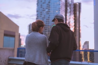 A couple stands on a rooftop or elevated urban area, with their backs to the camera. They are overlooking a cityscape with tall modern buildings and a partly cloudy sky. The woman is wearing a gray sweater, and the man is dressed in a black hoodie and a cap.