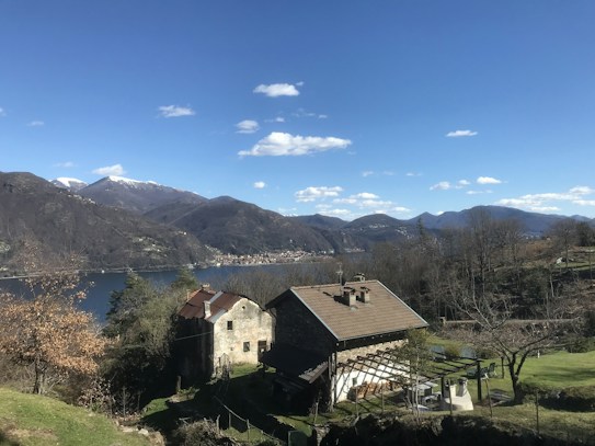A serene landscape features a rustic stone house with a pitched roof, nestled among trees on a hillside. In the background, a large lake stretches out, surrounded by mountains with patches of snow on their peaks. The sky is clear blue with a few scattered clouds.