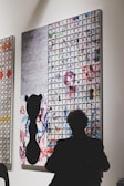 A person is silhouetted in front of an abstract art piece featuring a grid pattern with various colors and a black silhouetted shape in the foreground. The artwork combines geometric elements with splashes of color that appear haphazard and spontaneous.