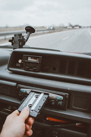 A hand is seen inserting a cassette tape into a car stereo on the dashboard of a vehicle. The road ahead is slightly blurred, emphasizing the focus on the dashboard, which includes various knobs and buttons.