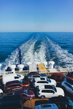 A ferry is transporting multiple parked cars across a vast expanse of blue ocean under a clear sky. The rear of the ferry shows the wake trailing behind, and numerous vehicles are tightly arranged on the ferry's deck.