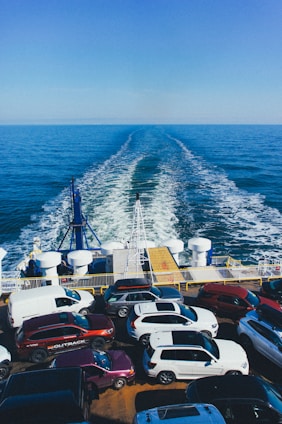 A ferry is transporting multiple parked cars across a vast expanse of blue ocean under a clear sky. The rear of the ferry shows the wake trailing behind, and numerous vehicles are tightly arranged on the ferry's deck.