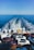A ferry is transporting multiple parked cars across a vast expanse of blue ocean under a clear sky. The rear of the ferry shows the wake trailing behind, and numerous vehicles are tightly arranged on the ferry's deck.