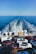 A ferry is transporting multiple parked cars across a vast expanse of blue ocean under a clear sky. The rear of the ferry shows the wake trailing behind, and numerous vehicles are tightly arranged on the ferry's deck.