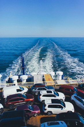 A ferry is transporting multiple parked cars across a vast expanse of blue ocean under a clear sky. The rear of the ferry shows the wake trailing behind, and numerous vehicles are tightly arranged on the ferry's deck.