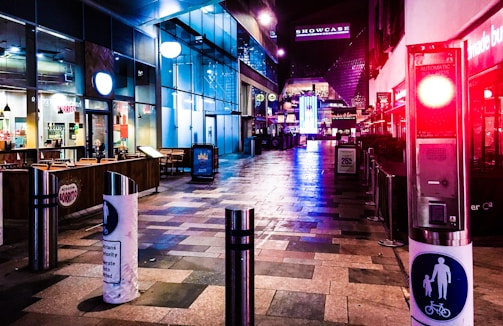 A modern outdoor shopping or entertainment area is illuminated with vibrant neon lights. The scene is empty, with storefronts along the sides and bright signage in the distance. The ground is composed of various colored tiles, and a couple of metallic bollards are visible in the foreground.