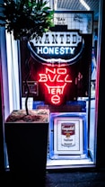 A neon sign with the words 'WANTED HONESTY NO BULL' is illuminated in a storefront. The sign features a bullhorn design with blue and red neon lights. Below the sign, there is a framed piece of art depicting a Campbell's Tartan Soup can. A potted plant with a leafy top is placed next to the framed art. The backdrop consists of a grid-like structure.