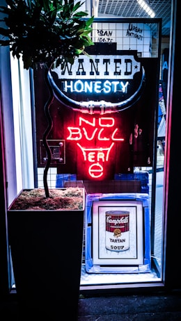 A neon sign with the words 'WANTED HONESTY NO BULL' is illuminated in a storefront. The sign features a bullhorn design with blue and red neon lights. Below the sign, there is a framed piece of art depicting a Campbell's Tartan Soup can. A potted plant with a leafy top is placed next to the framed art. The backdrop consists of a grid-like structure.