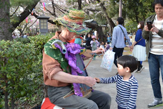 An elderly man wearing a colorful, knitted hat and vibrant layers of clothing is seated outdoors, interacting with a young child. The man appears to be performing, possibly playing a homemade instrument adorned with a purple and green puppet. The child, dressed in a striped shirt, is attentively engaging with him. People are walking and standing around, suggesting a park or festival setting, with trees and a lantern visible in the background.