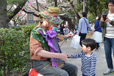 An elderly man wearing a colorful, knitted hat and vibrant layers of clothing is seated outdoors, interacting with a young child. The man appears to be performing, possibly playing a homemade instrument adorned with a purple and green puppet. The child, dressed in a striped shirt, is attentively engaging with him. People are walking and standing around, suggesting a park or festival setting, with trees and a lantern visible in the background.