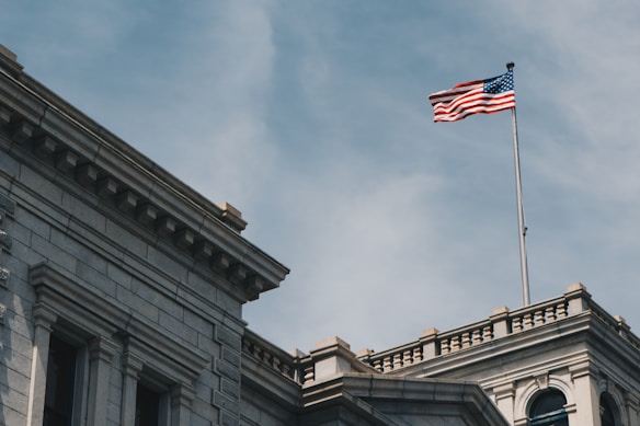A beige stone building with classical architecture, featuring columns and decorative cornices. An American flag is prominently displayed on a tall flagpole, waving against a clear blue sky.