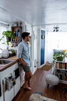 A person is casually leaning against a white kitchen counter in a small, cozy living space. The room has wooden flooring and is decorated with green plants. A bookshelf is filled with books and decorative items. A small footstool with patterned fabric is in the foreground. Natural light floods the room through sheer curtains, creating a bright and airy atmosphere.