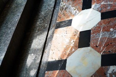 Geometric tiles are arranged in an intricate pattern with octagonal and square shapes made of marble, featuring a combination of white, black, and reddish colors. Sunlight casts shadows across the surface, creating a play of light and shadow that highlights the texture of the materials.