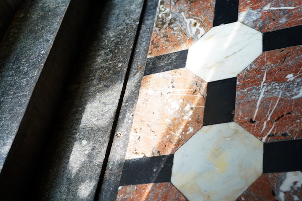 Geometric tiles are arranged in an intricate pattern with octagonal and square shapes made of marble, featuring a combination of white, black, and reddish colors. Sunlight casts shadows across the surface, creating a play of light and shadow that highlights the texture of the materials.