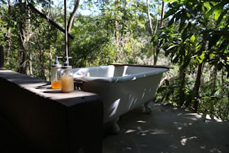 A vintage-style white bathtub is placed outdoors amidst lush greenery. The setting is peaceful and natural with sunlight filtering through the leaves. Two glass bottles with pump dispensers, one containing a yellowish liquid and the other a clear amber liquid, sit on a concrete ledge nearby.