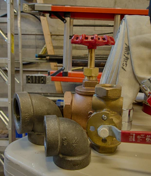 A collection of plumbing fittings and components is arranged on a surface. Notable items include a brass valve with a red handle, a couple of elbow joints, and a rolled-up white hose. In the background, a red and black ladder can be seen along with some metal poles against a concrete wall.