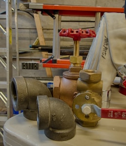 A collection of plumbing fittings and components is arranged on a surface. Notable items include a brass valve with a red handle, a couple of elbow joints, and a rolled-up white hose. In the background, a red and black ladder can be seen along with some metal poles against a concrete wall.