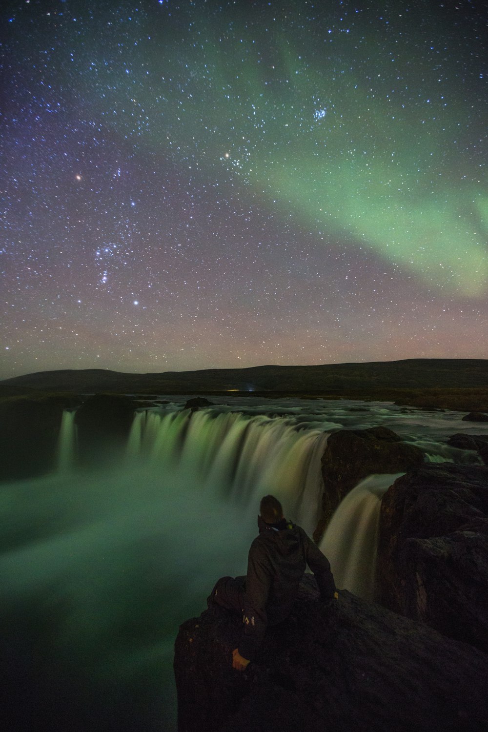 person sitting near waterfalls under aurora lights