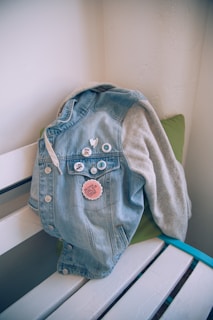 A denim jacket with several pins attached is draped over a white wooden bench. A green cushion provides a subtle contrast to the jacket, while textured white walls form the background.