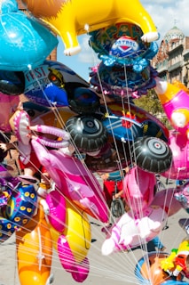 A cluster of colorful inflatable balloons, shaped like animals and cartoon characters, held together by strings. The background shows a street scene with buildings and people moving around.