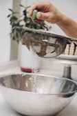 A hand is holding a metal sieve with a green handle, sifting white powder into a large stainless steel bowl. The action is taking place in a kitchen setting, with a blurred plant in the background.