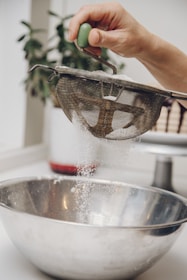 A hand is holding a metal sieve with a green handle, sifting white powder into a large stainless steel bowl. The action is taking place in a kitchen setting, with a blurred plant in the background.