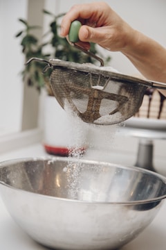 A hand is holding a metal sieve with a green handle, sifting white powder into a large stainless steel bowl. The action is taking place in a kitchen setting, with a blurred plant in the background.