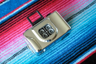 A vintage-style camera with multiple lenses and a metallic finish sits on a colorful striped fabric. The fabric features vibrant shades of blue, pink, and red with black lines adding contrast.