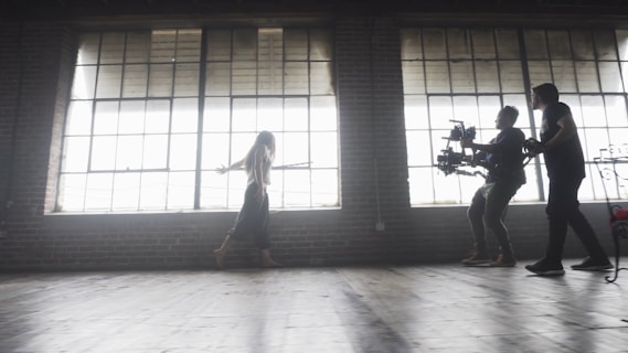 A dimly lit rustic studio features large, industrial-style windows casting soft light across the wooden floor. A woman walks gracefully, arms outstretched, while two individuals operate a professional camera rig, capturing her movement.