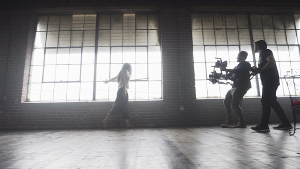 A dimly lit rustic studio features large, industrial-style windows casting soft light across the wooden floor. A woman walks gracefully, arms outstretched, while two individuals operate a professional camera rig, capturing her movement.