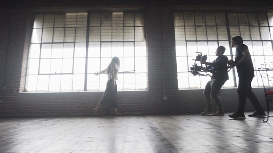 A dimly lit rustic studio features large, industrial-style windows casting soft light across the wooden floor. A woman walks gracefully, arms outstretched, while two individuals operate a professional camera rig, capturing her movement.