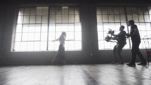 A dimly lit rustic studio features large, industrial-style windows casting soft light across the wooden floor. A woman walks gracefully, arms outstretched, while two individuals operate a professional camera rig, capturing her movement.