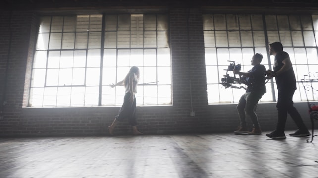 A dimly lit rustic studio features large, industrial-style windows casting soft light across the wooden floor. A woman walks gracefully, arms outstretched, while two individuals operate a professional camera rig, capturing her movement.