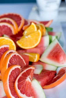 Slices of various fruits including grapefruit, orange, and watermelon are arranged on a white surface. The fruits are fresh and colorful, showing their vibrant red, orange, and green hues. A blurred background includes what appears to be a cup.