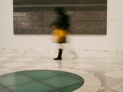 A blurred image of a person in motion appears in front of a large, textured artwork in a gallery setting. The floor is tiled with a pattern of squares, and there is a circular design featuring shades of green. The person appears to be wearing a yellow garment and black boots.