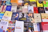 A variety of books are displayed on a table. The collection includes titles in both English and Chinese, and covers multiple genres. Prominent cover images include a man in a suit, a classic film scene, and abstract artwork. Many of the books have bright, colorful covers, while others are more muted.