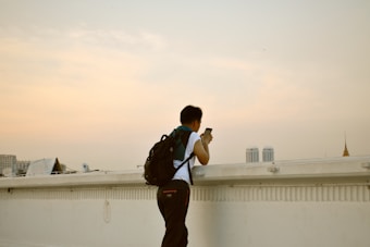 A person with a backpack is standing on a rooftop, looking towards the horizon and holding a smartphone. The sky is overcast with gentle shades of grey and orange, and a cityscape with various buildings is visible in the background.