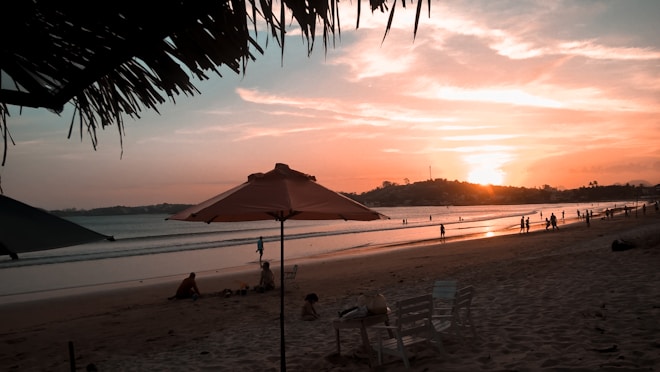 The beach scene captures a serene sunset with the sun setting behind distant hills. The sky is filled with warm hues of orange and pink. People are leisurely walking and enjoying the shoreline, while beach chairs and umbrellas are set up in the sand. Palm leaves frame the top of the image, adding a tropical feel.