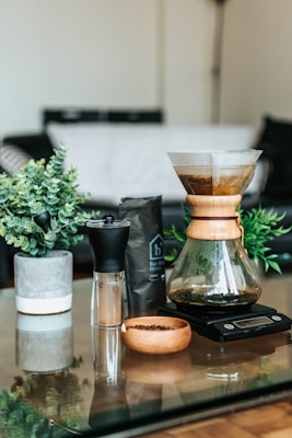 A coffee brewing setup is placed on a glass table, featuring a Chemex-style coffee maker on a digital scale, a wooden bowl with coffee beans, a manual coffee grinder with a black top, and a bag of coffee. There are two small potted plants for decoration, and the background shows a blurred living room setting with a couch.