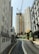 A construction site is situated between tall, residential high-rise buildings. The focus is on a tall, yellow construction crane towering over the site, surrounded by green scaffolding and construction barriers. The road curves towards the construction site, and a few air conditioning units are visible on the sides of the nearby buildings.