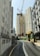 A construction site is situated between tall, residential high-rise buildings. The focus is on a tall, yellow construction crane towering over the site, surrounded by green scaffolding and construction barriers. The road curves towards the construction site, and a few air conditioning units are visible on the sides of the nearby buildings.