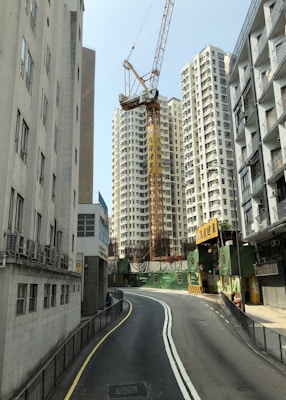 A construction site is situated between tall, residential high-rise buildings. The focus is on a tall, yellow construction crane towering over the site, surrounded by green scaffolding and construction barriers. The road curves towards the construction site, and a few air conditioning units are visible on the sides of the nearby buildings.
