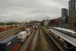 A bustling railway and shipping container yard set against an urban backdrop of tall buildings. Numerous colorful shipping containers are aligned on multiple train tracks, while a passenger train is stationed at a nearby platform. The sky is overcast, adding a dramatic tone to the scene.