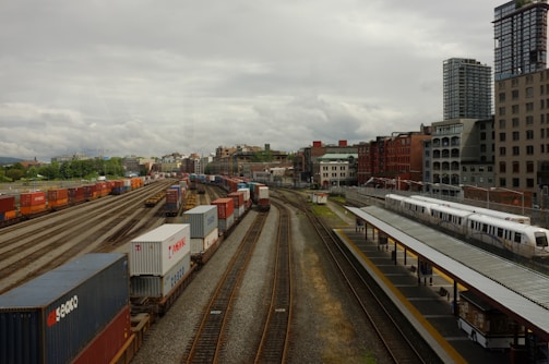 A bustling railway and shipping container yard set against an urban backdrop of tall buildings. Numerous colorful shipping containers are aligned on multiple train tracks, while a passenger train is stationed at a nearby platform. The sky is overcast, adding a dramatic tone to the scene.